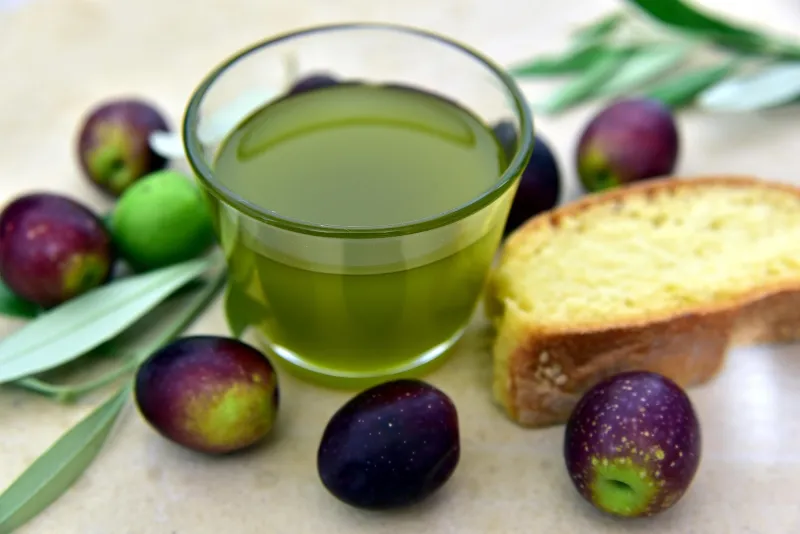 Un vaso de aceite verde entre aceitunas, pan y ramas de olivo
